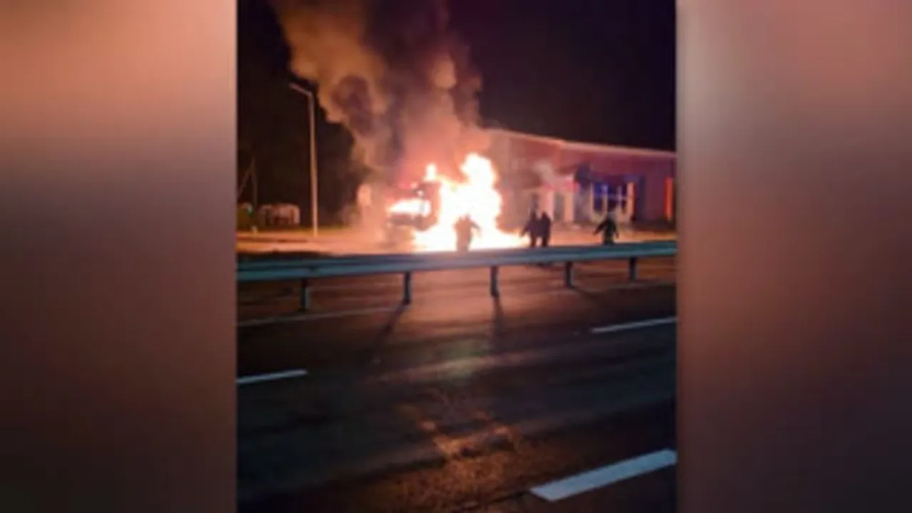 Vehículo en llamas de noche cerca de una estructura con personas y humo espeso tras un impacto.