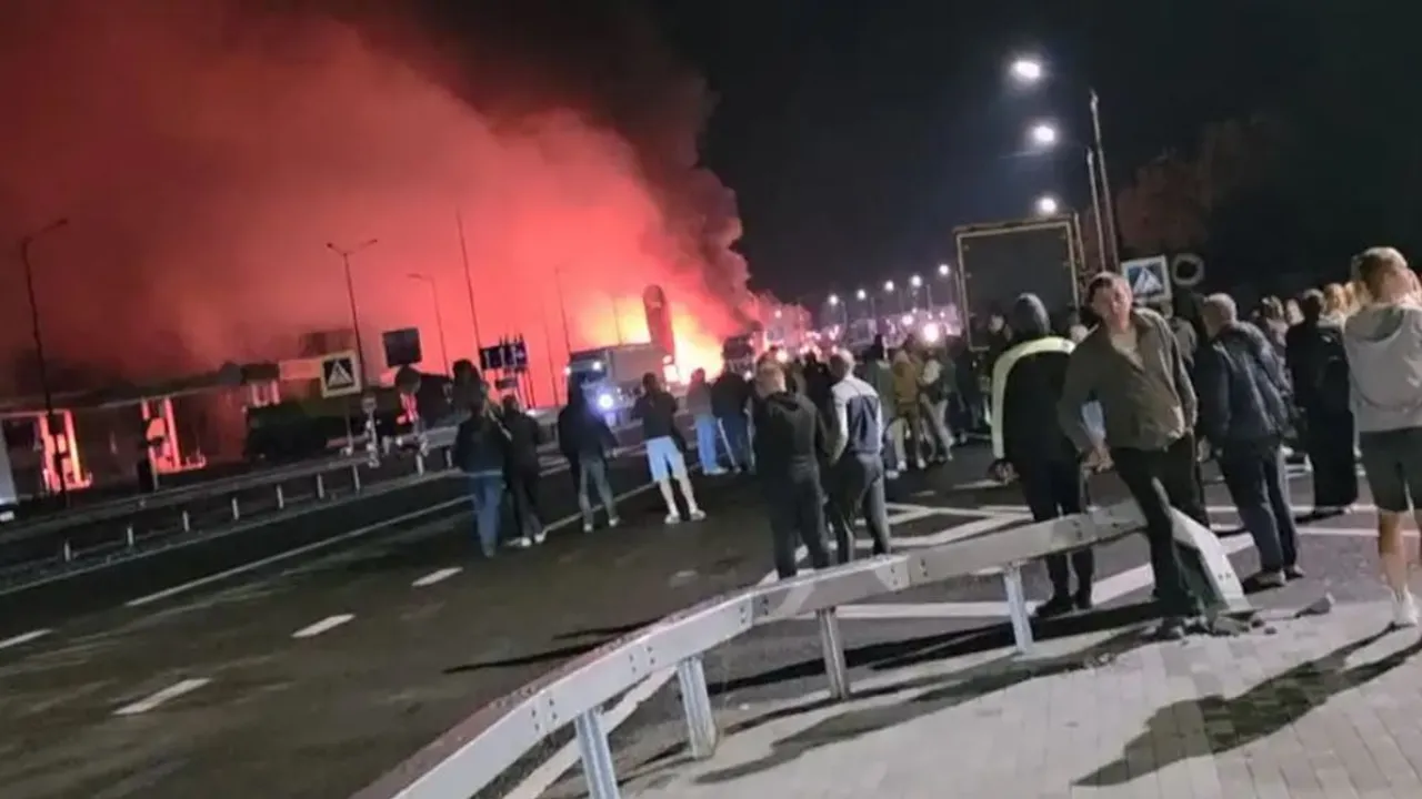Personas reunidas de noche en una carretera observan a lo lejos un gran incendio con columnas de humo y resplandor rojo.