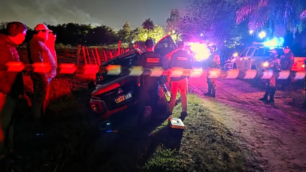 Camioneta volcada en una zanja al costado de un camino de tierra resguardada por policías con cinta perimetral de noche.