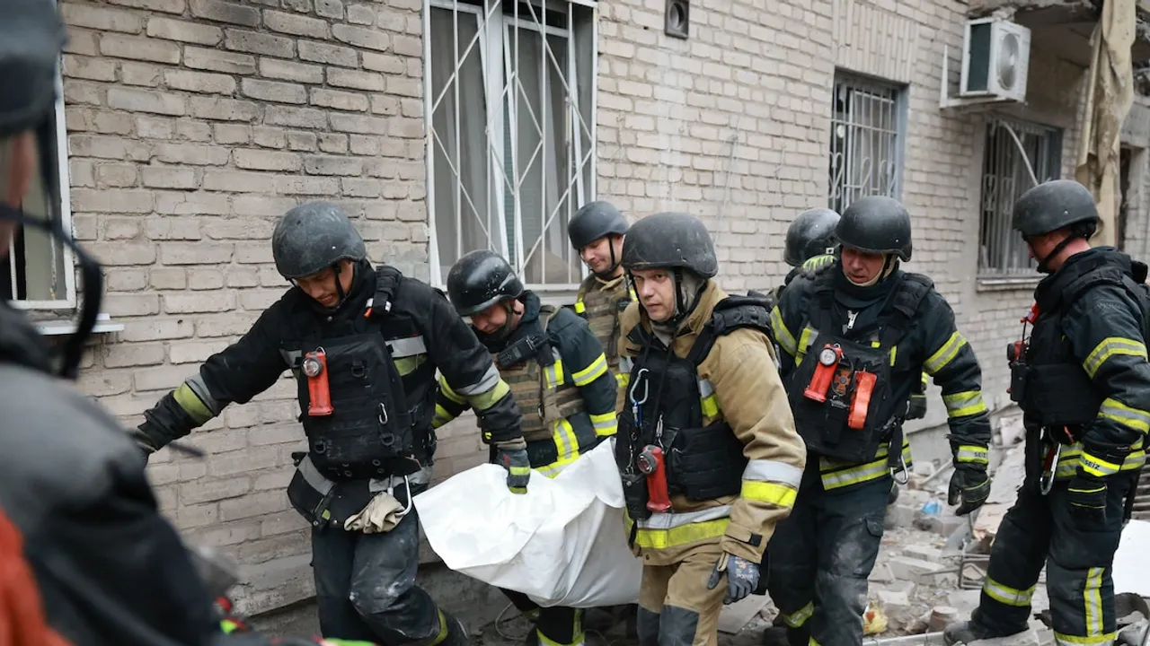 Bomberos con cascos y chalecos trasladan en una camilla cubierta a una víctima frente a un edificio dañado.