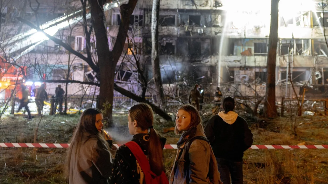 Personas observan un edificio residencial dañado de noche tras un bombardeo mientras bomberos trabajan en el lugar.