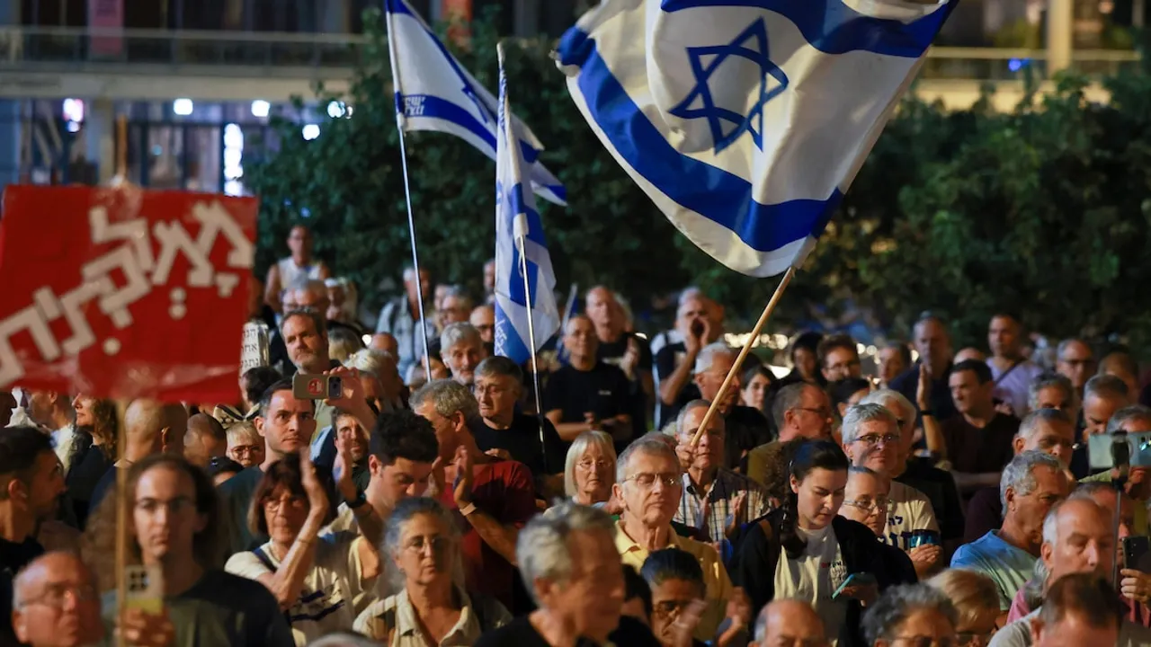 Multitud reunida durante la noche sostiene pancartas y una gran bandera de Israel con la estrella de David flameando.