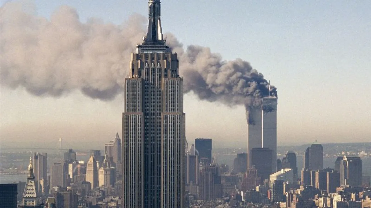 El edificio Empire State en primer plano con las Torres Gemelas humeando al fondo bajo un cielo claro.