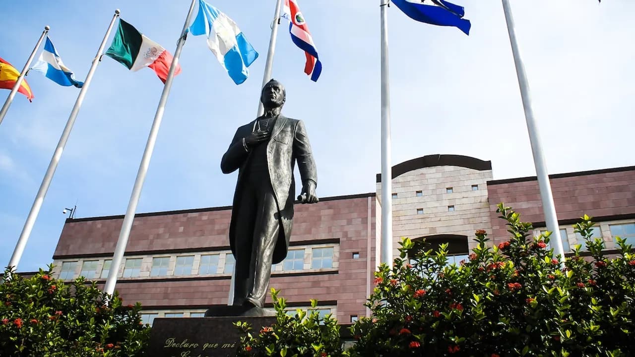 Estatua de bronce de prócer frente a edificio institucional rodeada de banderas latinoamericanas ondeando al viento.