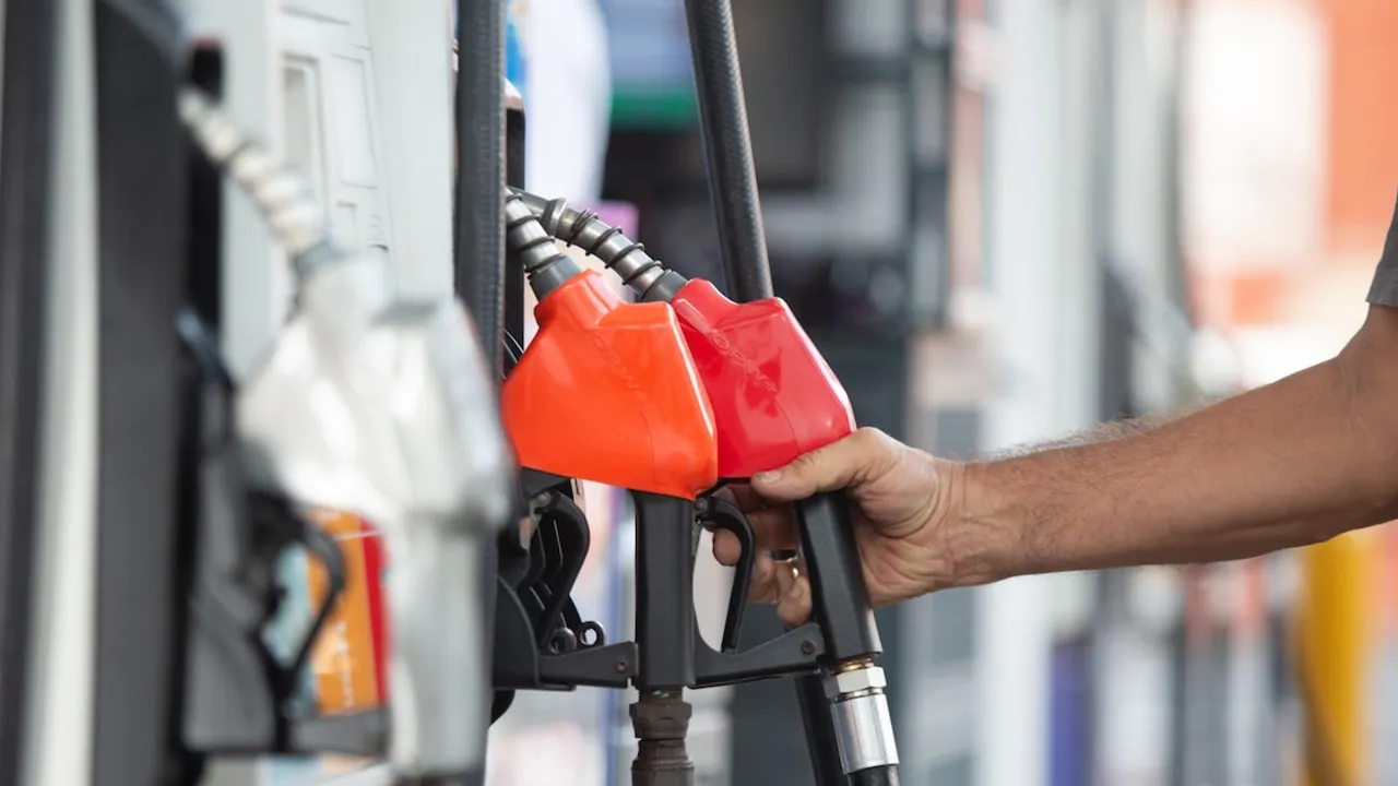 Mano sosteniendo una pistola surtidora roja de combustible en una estación de servicio junto a otra manguera naranja.