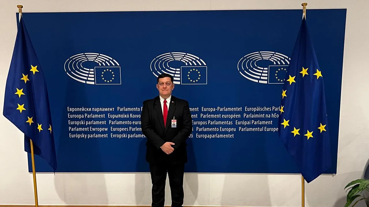 Hombre de traje oscuro y corbata roja posa frente a un panel azul y dos banderas de la Unión Europea.