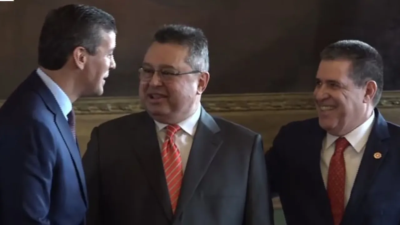 Tres hombres con traje y corbata conversan sonrientes en un salón institucional durante un evento oficial.