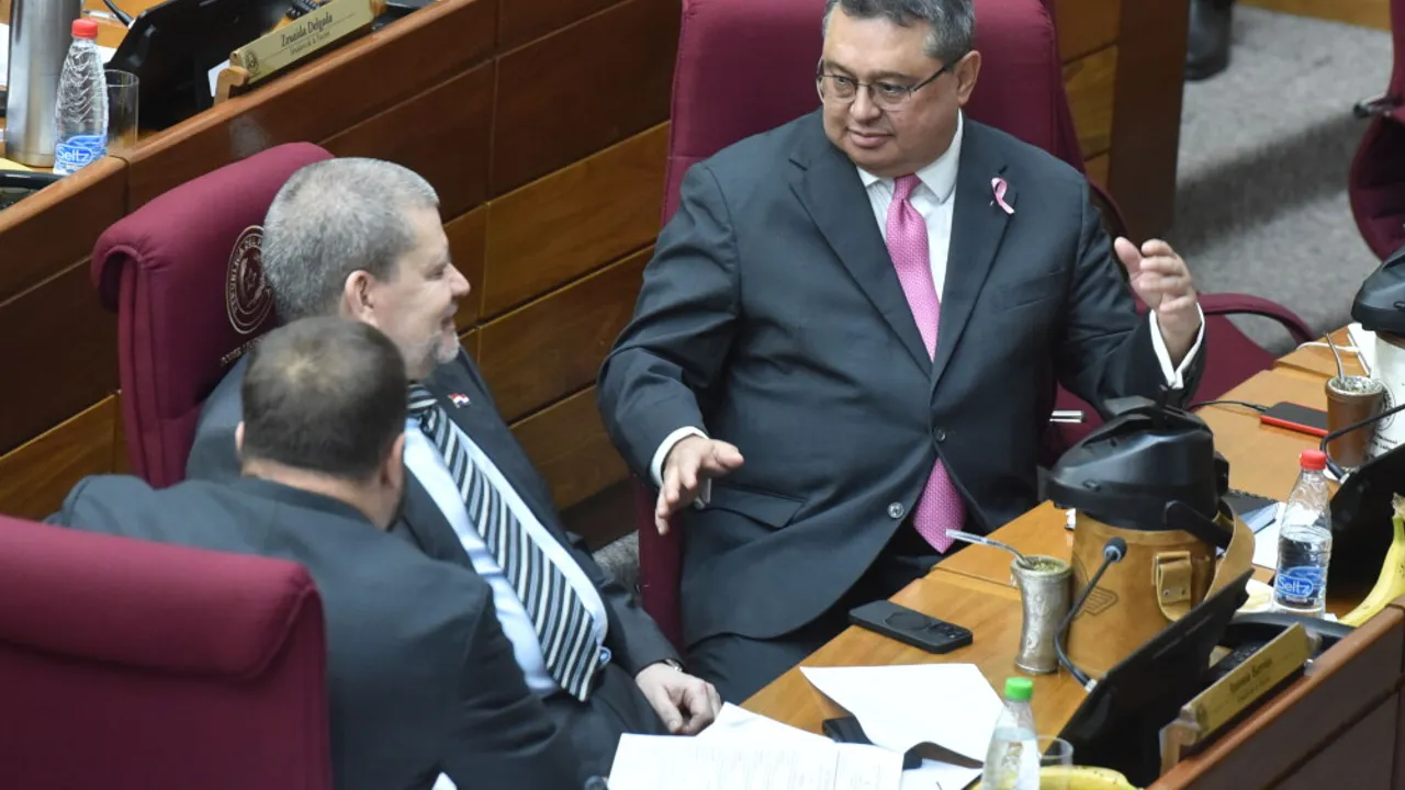 Dos senadores vestidos con traje oscuro conversan sentados en sus bancas de la Cámara de Senadores de Paraguay.