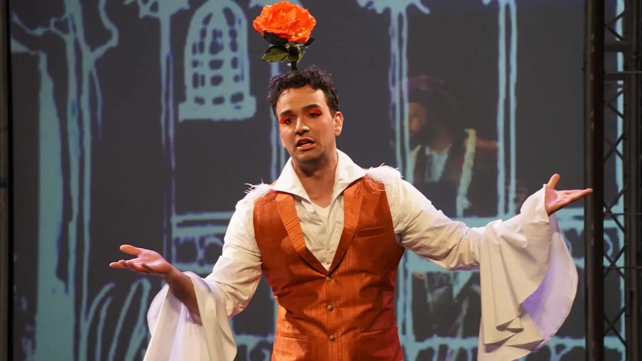 Hombre caracterizado para ópera con una flor naranja en la cabeza y chaleco marrón con los brazos extendidos