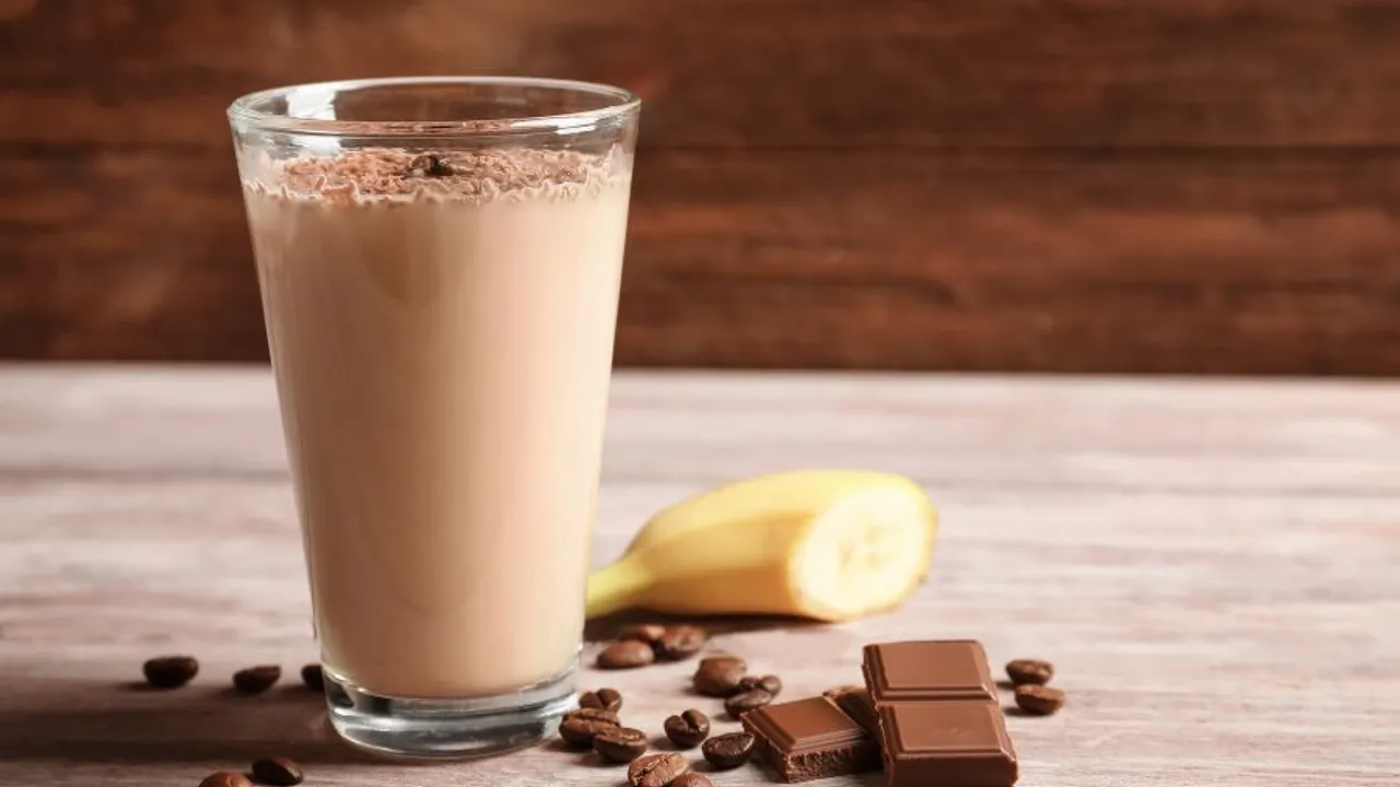 Vaso con batido de proteínas sobre mesa de madera junto a granos de café, chocolate y banana.