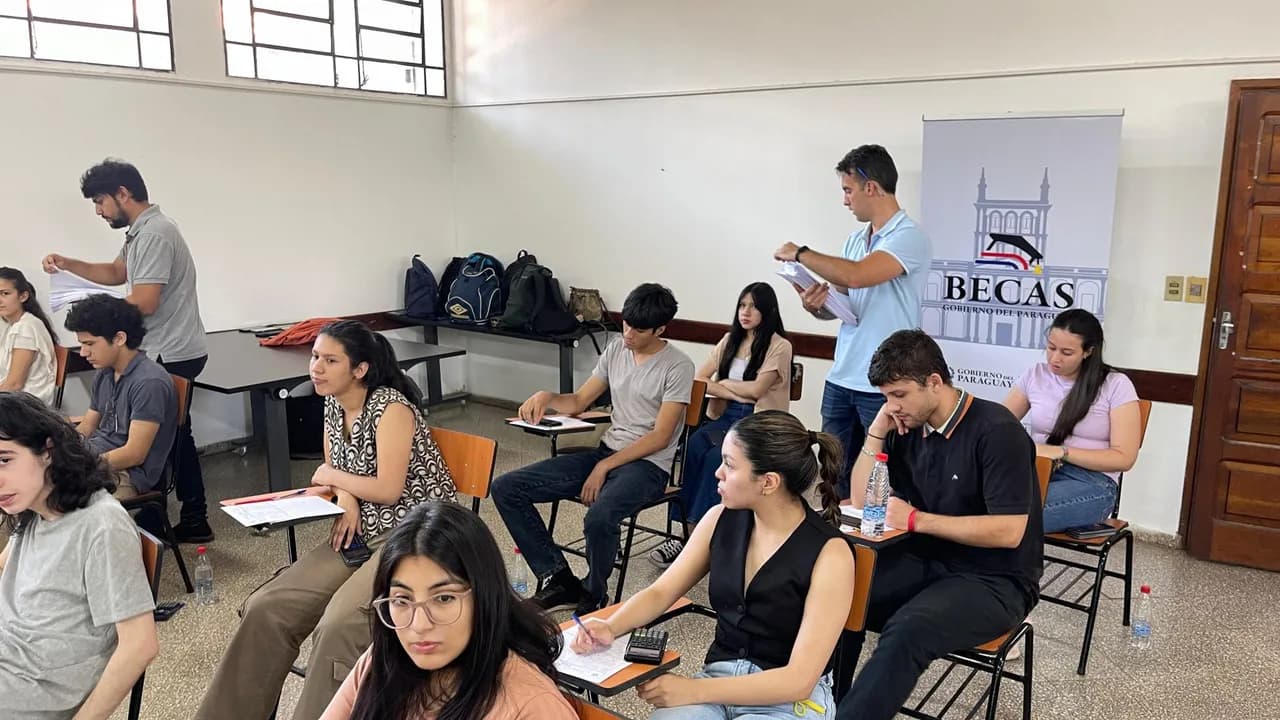 Jóvenes sentados en un aula completan formularios mientras un evaluador distribuye hojas frente a un cartel.