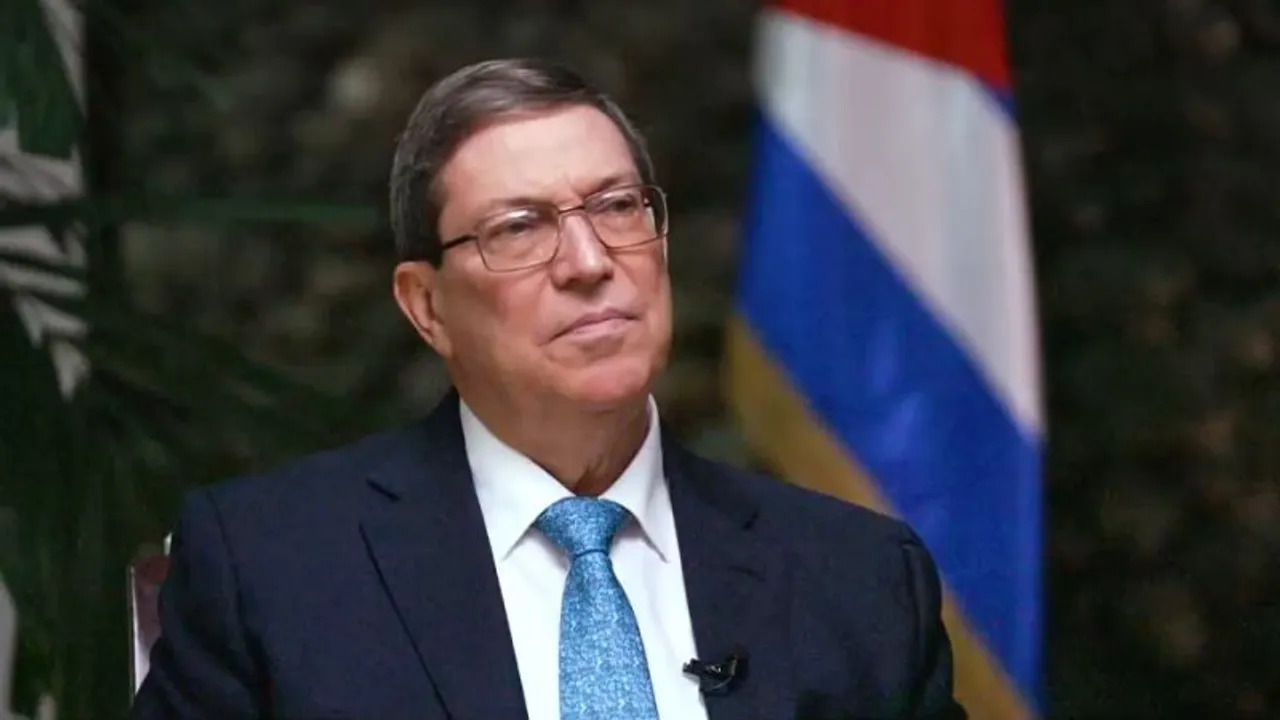 Retrato del canciller cubano Bruno Rodríguez con traje oscuro y corbata azul, con la bandera de Cuba detrás.