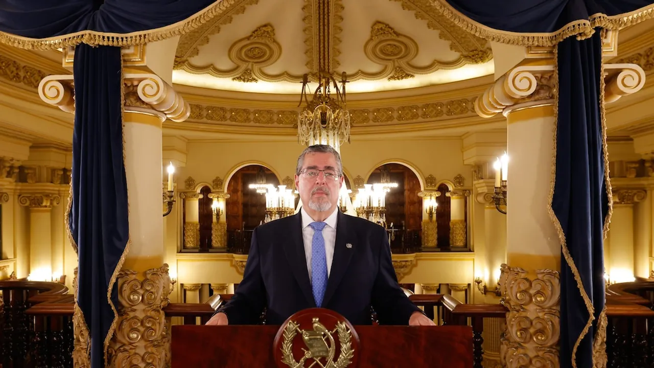 Bernardo Arévalo con traje oscuro y corbata azul habla detrás de un podio en un salón con columnas doradas.