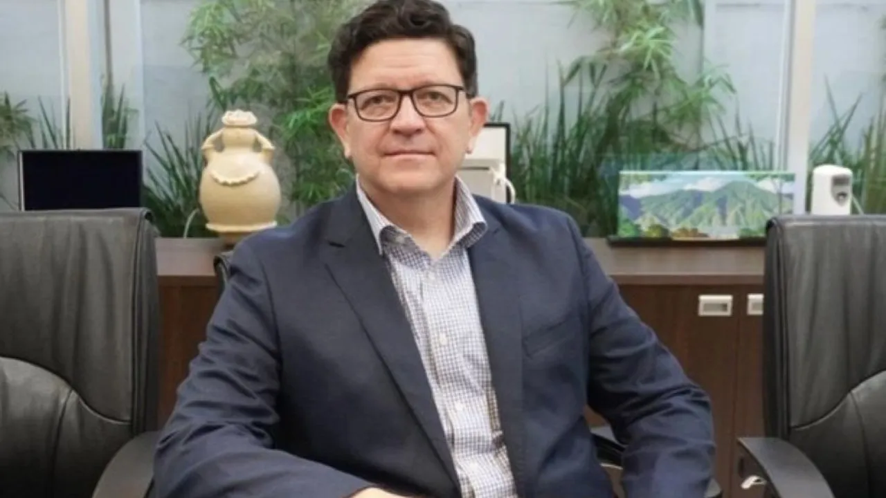 Hombre con anteojos y saco oscuro sentado en una sala de reuniones con fondo de plantas y elementos decorativos.