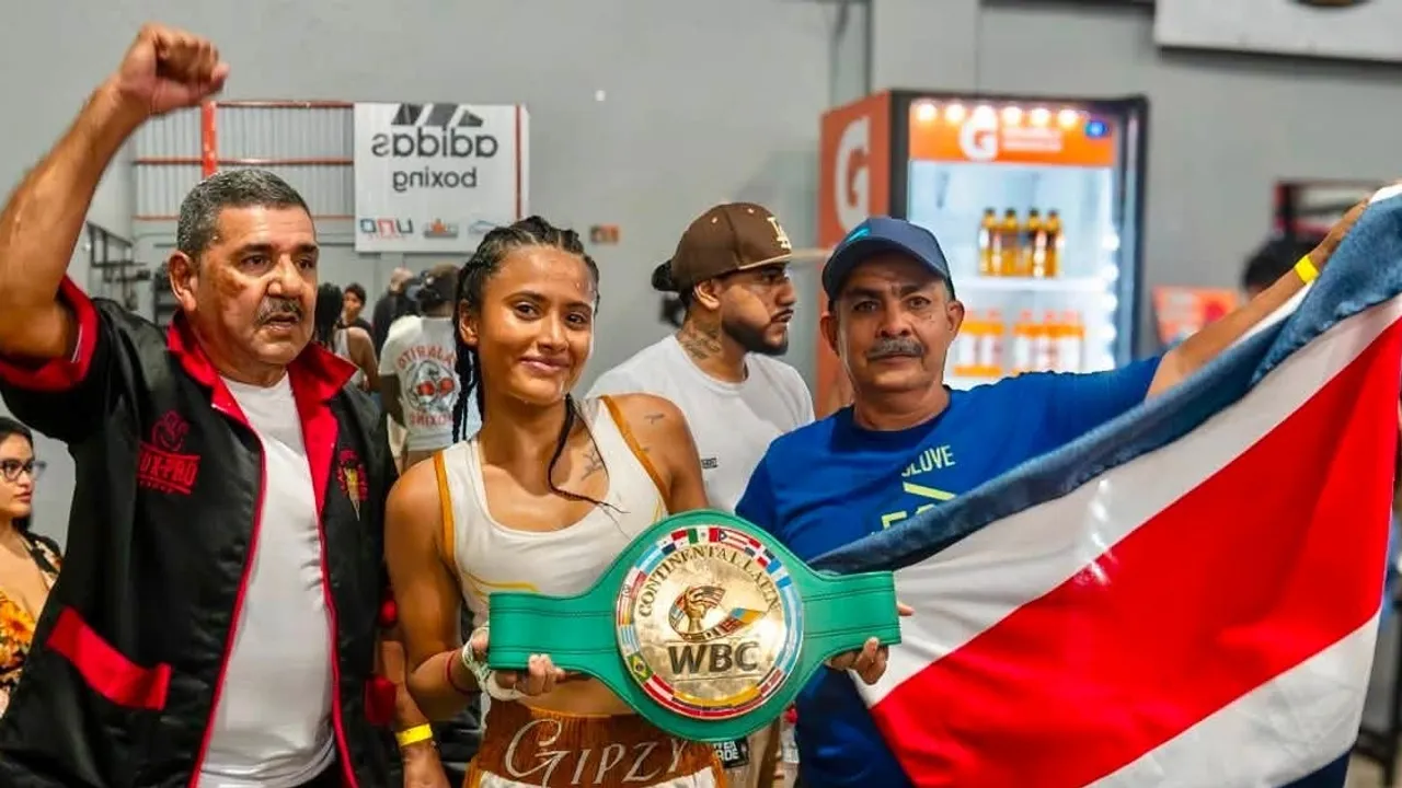 Pugilista sonriente sosteniendo un cinturón verde de boxeo junto a dos entrenadores y la bandera costarricense.