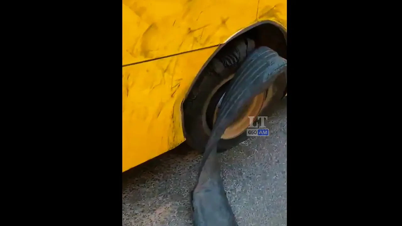 Rueda trasera averiada de un colectivo amarillo con la cubierta de goma desprendida sobre el asfalto.