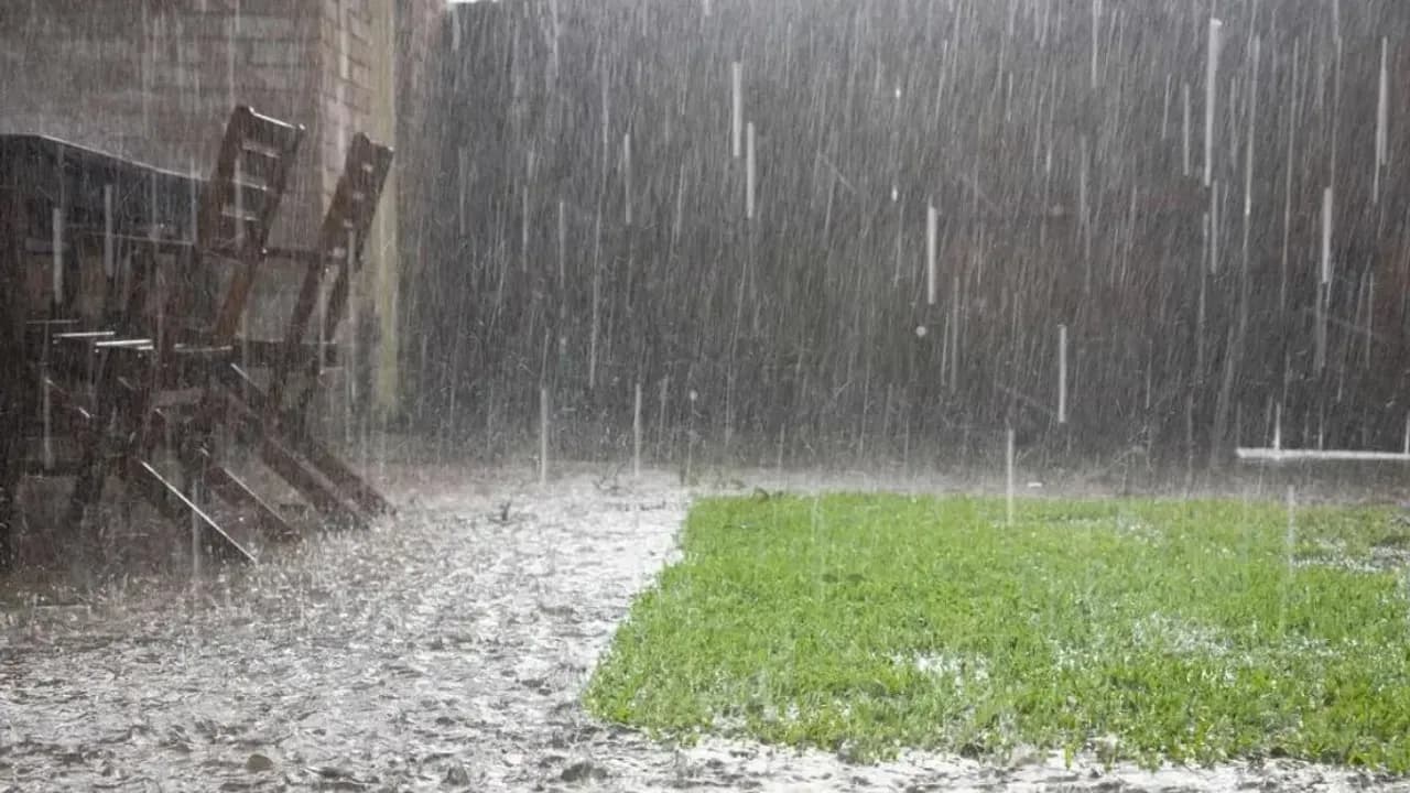 Lluvia intensa cayendo sobre un patio con césped verde y sillas de madera al aire libre durante un fuerte temporal.