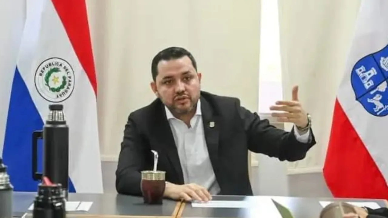 Hombre con saco oscuro y camisa blanca habla en una mesa de reuniones frente a una guampa y dos banderas oficiales.