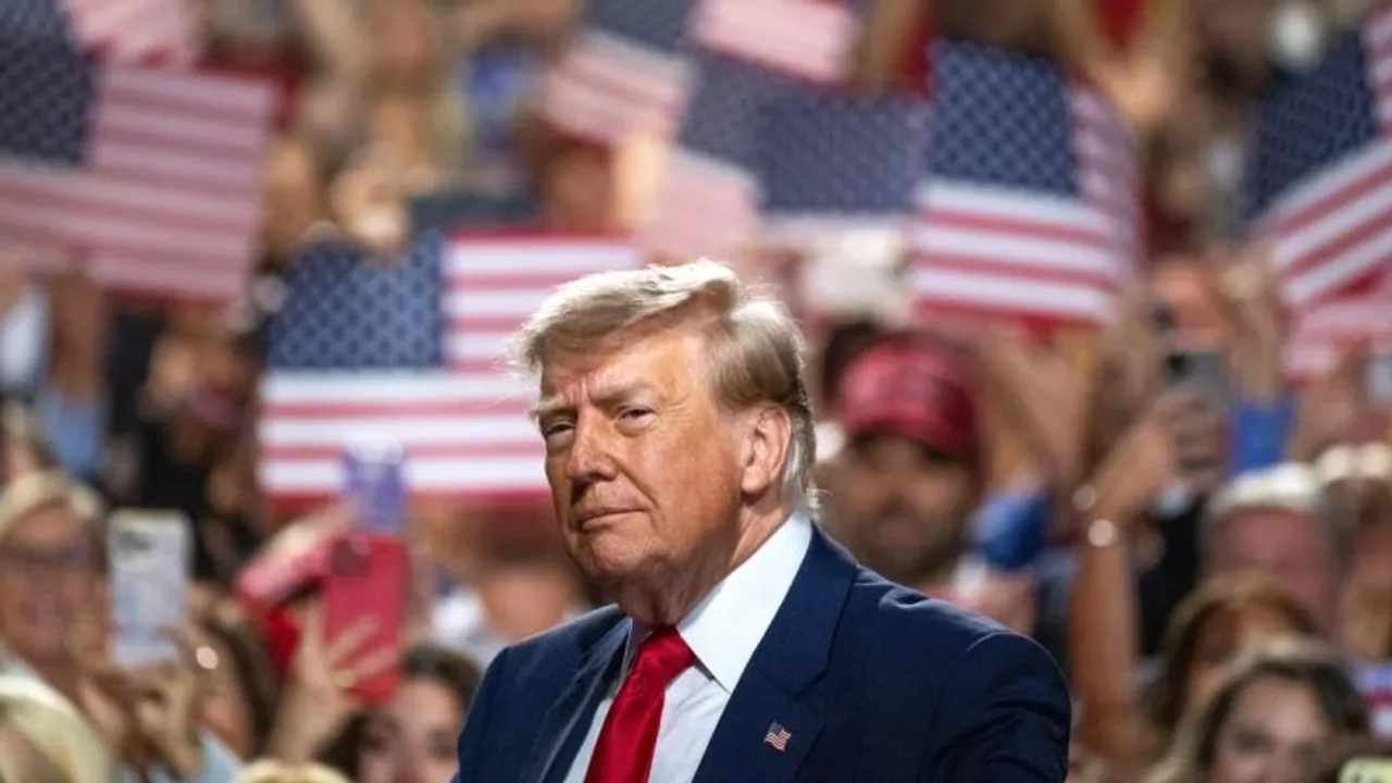 Donald Trump con traje azul y corbata roja frente a una multitud que sostiene banderas de Estados Unidos.