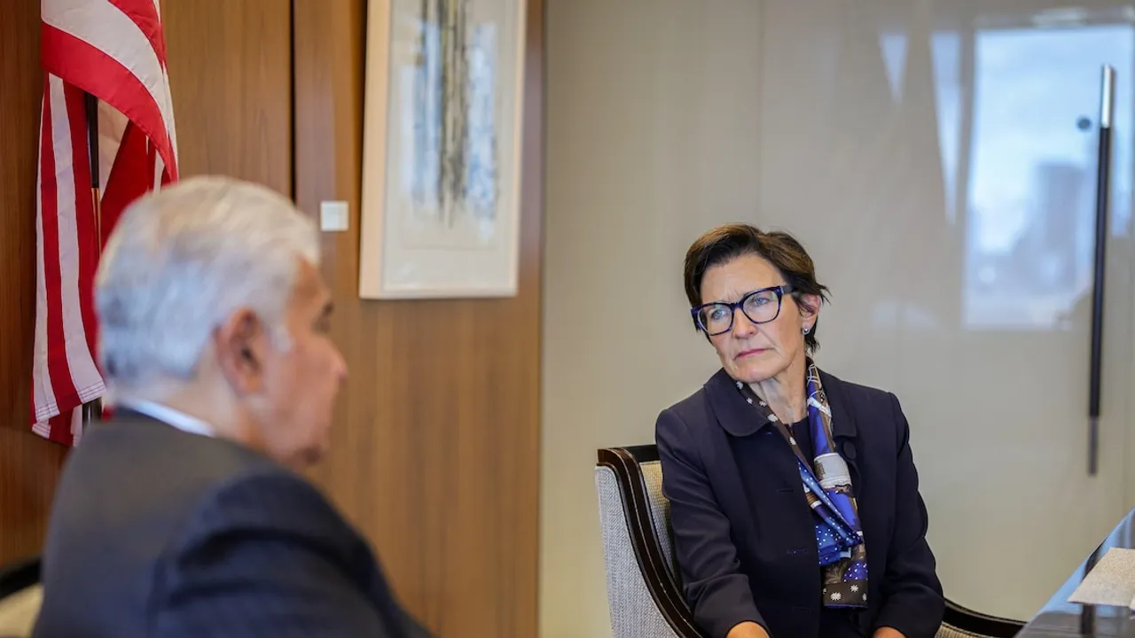Jane Fraser sentada con anteojos y saco oscuro dialoga en una reunión corporativa frente a una bandera estadounidense.