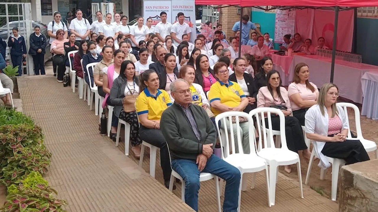 Personas sentadas en sillas plásticas al aire libre durante una charla médica comunitaria frente a una carpa rosada.