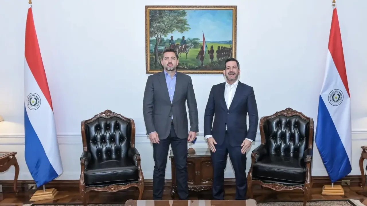 Pedro Alliana y Raúl Latorre posan de pie en una oficina entre dos banderas paraguayas y un cuadro histórico.