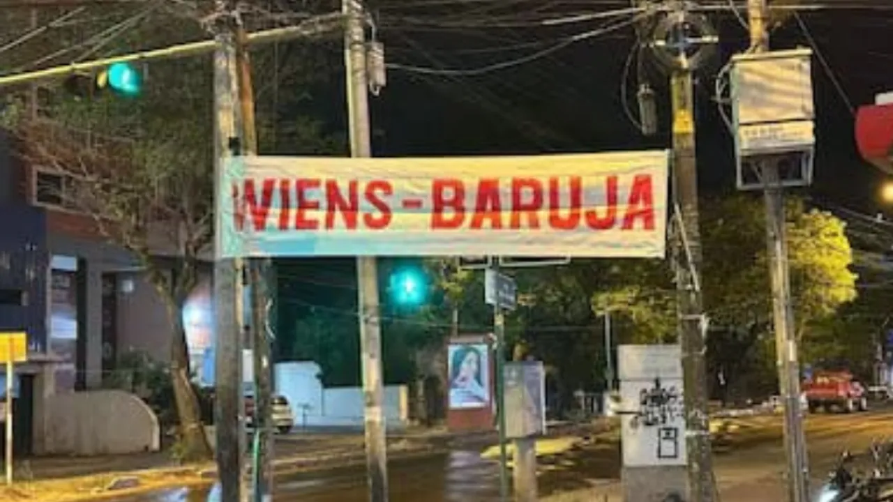 Pasacalle con la inscripción Wiens Baruja colgado entre postes de tendido eléctrico sobre una calle nocturna.