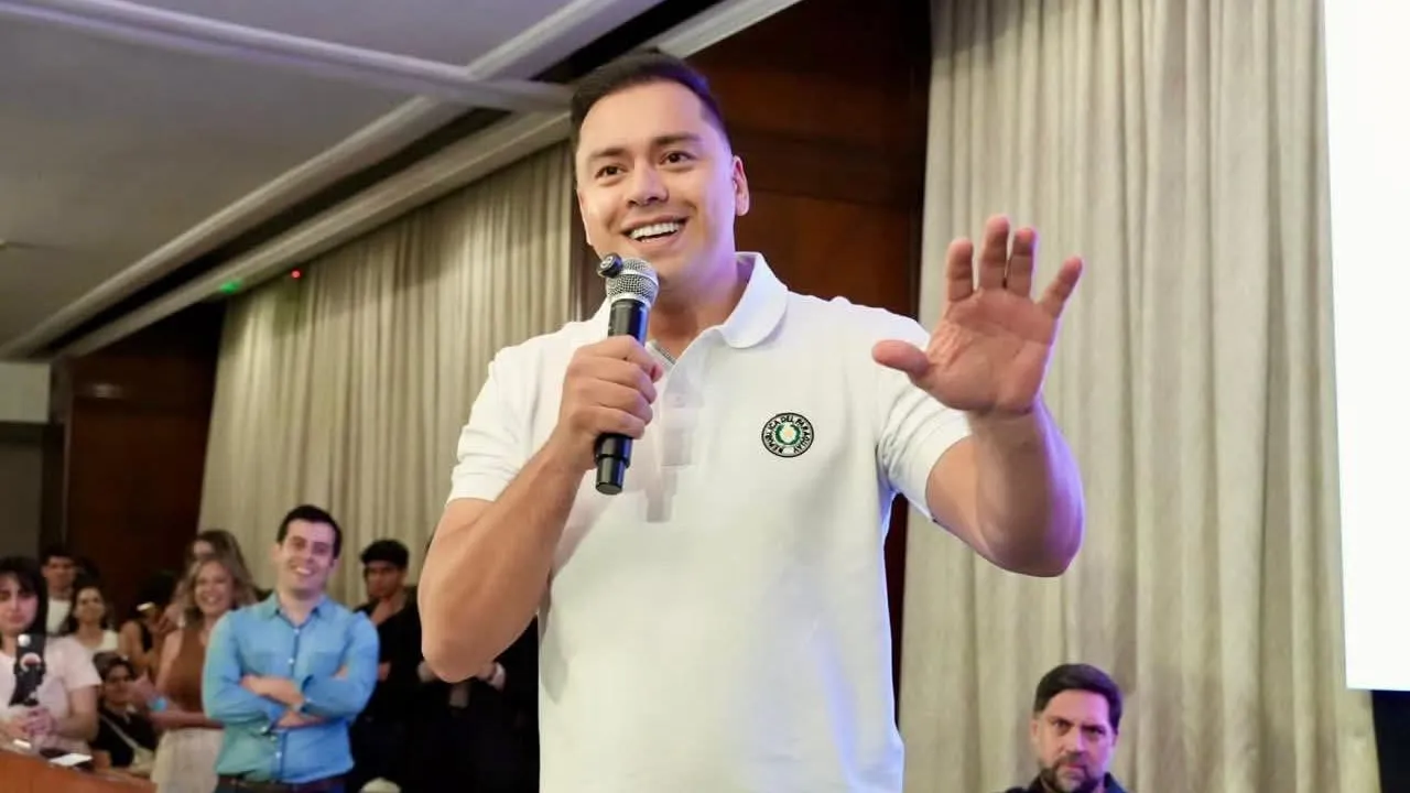 Hombre con remera blanca y micrófono en mano habla sonriente ante un auditorio durante un encuentro político.