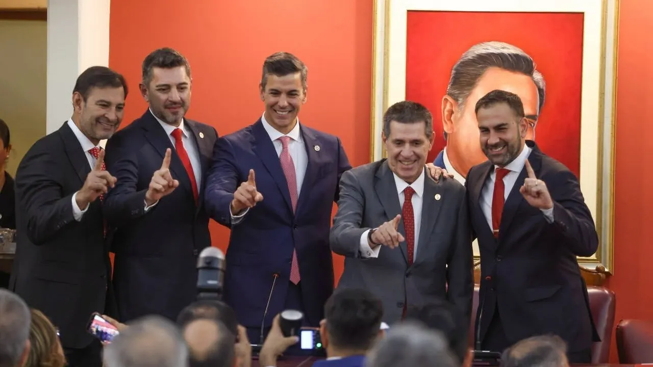 Cinco dirigentes políticos en traje saludan sonrientes en un salón oficial delante de un cuadro con retrato.
