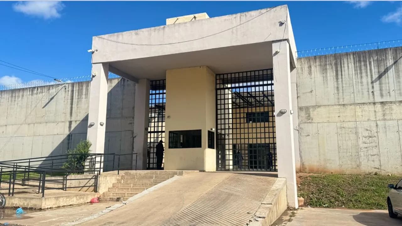 Entrada principal de una cárcel con rejas negras, altos muros de cemento y alambre de púas perimetral.