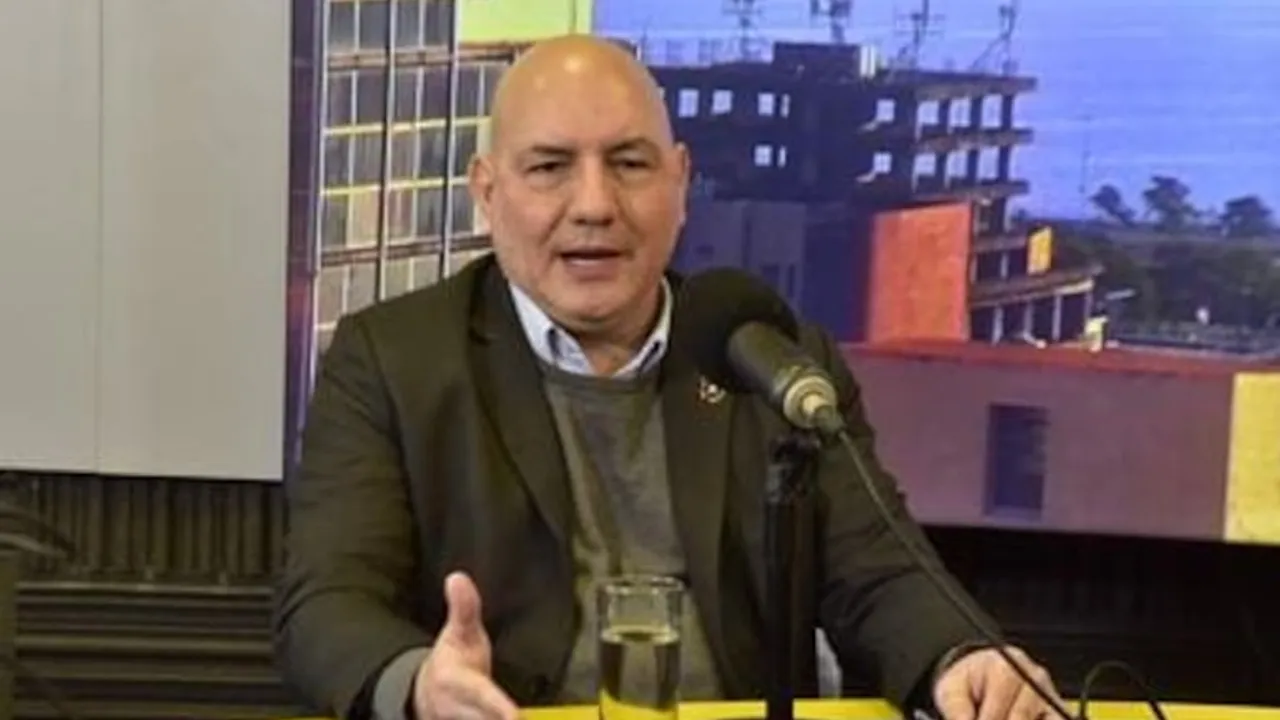 Hombre calvo con traje oscuro y suéter gris hablando frente a un micrófono en un estudio televisivo.