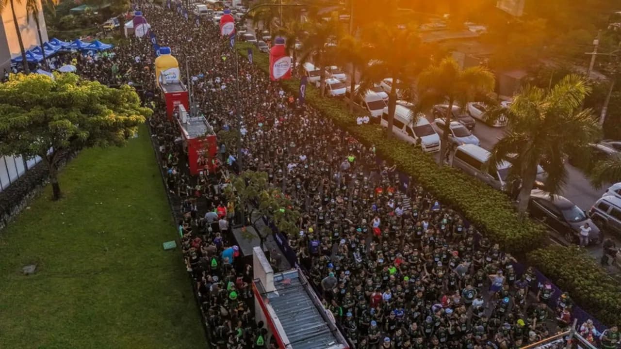 Multitud de personas corriendo en una avenida urbana al amanecer, flanqueada por vehículos y vegetación.