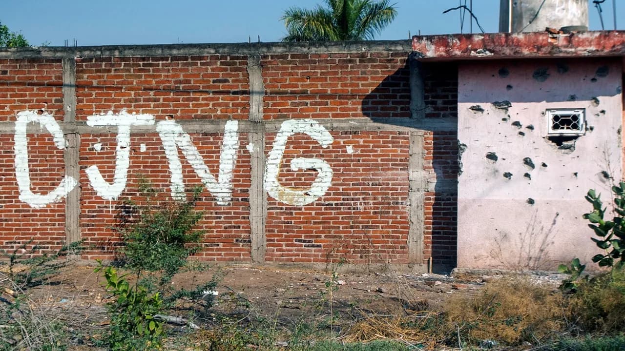 Muro de ladrillos visto con la sigla CJNG pintada en blanco y una pared rosada contigua con marcas de disparos.