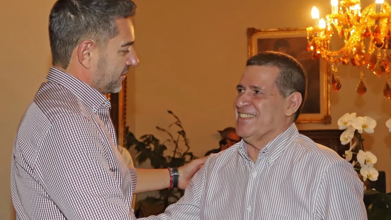 Pedro Alliana y Horacio Cartes conversan sonrientes en un salón iluminado con una araña clásica de luces.