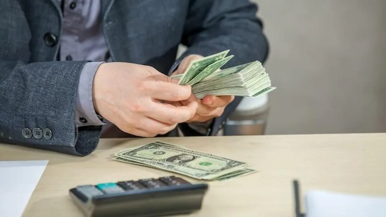 Persona en traje cuenta fajos de billetes de dólares estadounidenses sobre un escritorio junto a una calculadora.