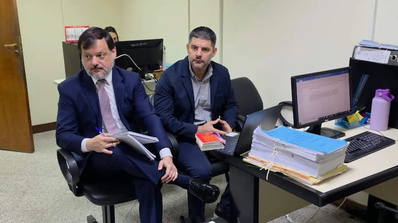 Dos hombres en traje sentados ante un escritorio de oficina con carpetas de expedientes judiciales y una computadora.