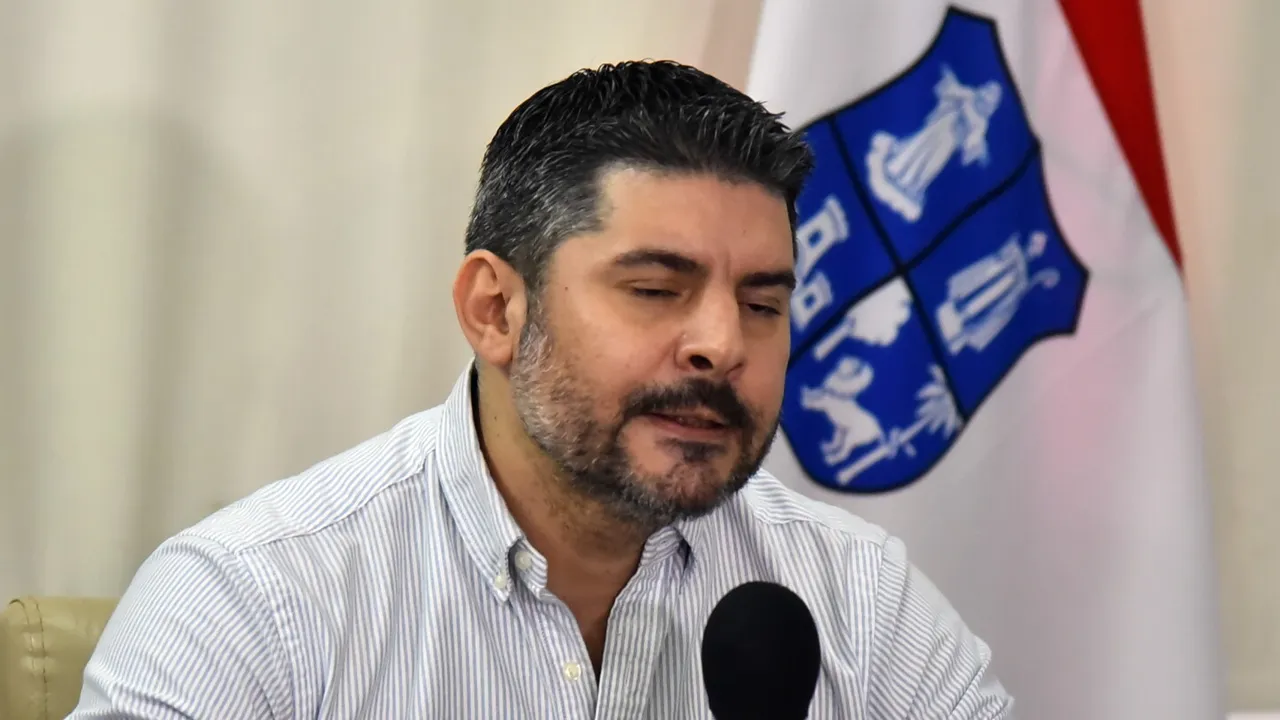 Óscar Rodríguez habla frente a un micrófono con camisa a rayas y un escudo institucional de fondo