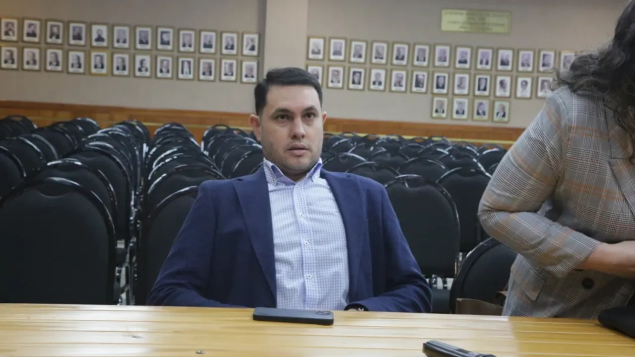Hernán Rivas sentado con saco azul y camisa a cuadros en una sala del Poder Judicial con sillas vacías al fondo.