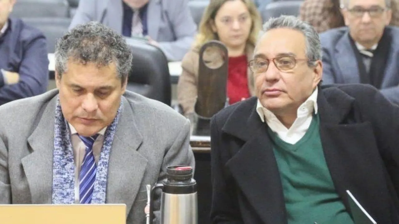 Dos hombres con abrigos oscuros sentados en bancas durante un acto público o sesión parlamentaria