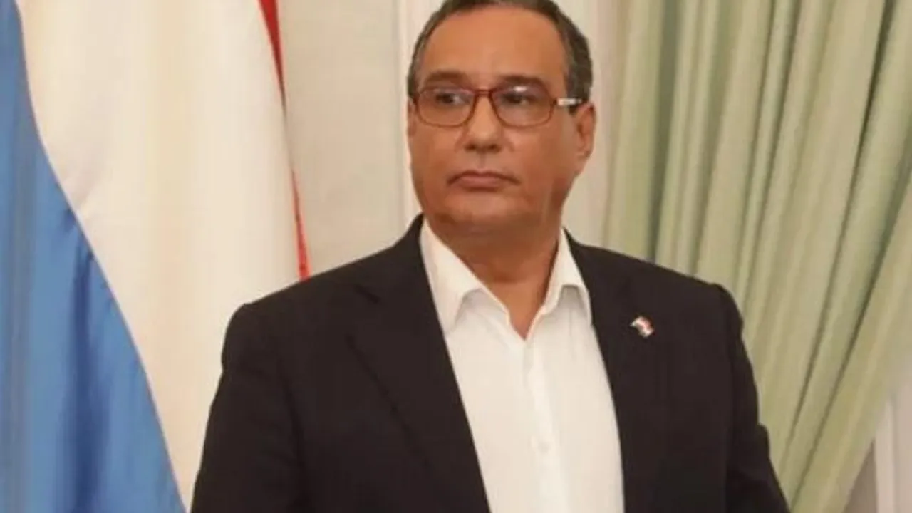 Hombre de traje oscuro, camisa blanca y anteojos frente a una bandera paraguaya.