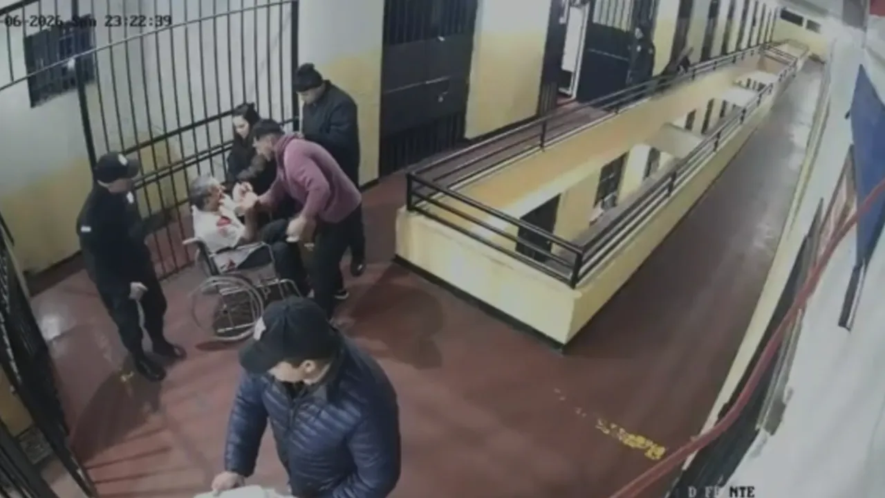 Cámara de seguridad penitenciaria muestra a guardias y reclusos asistiendo a un interno sentado en silla de ruedas.