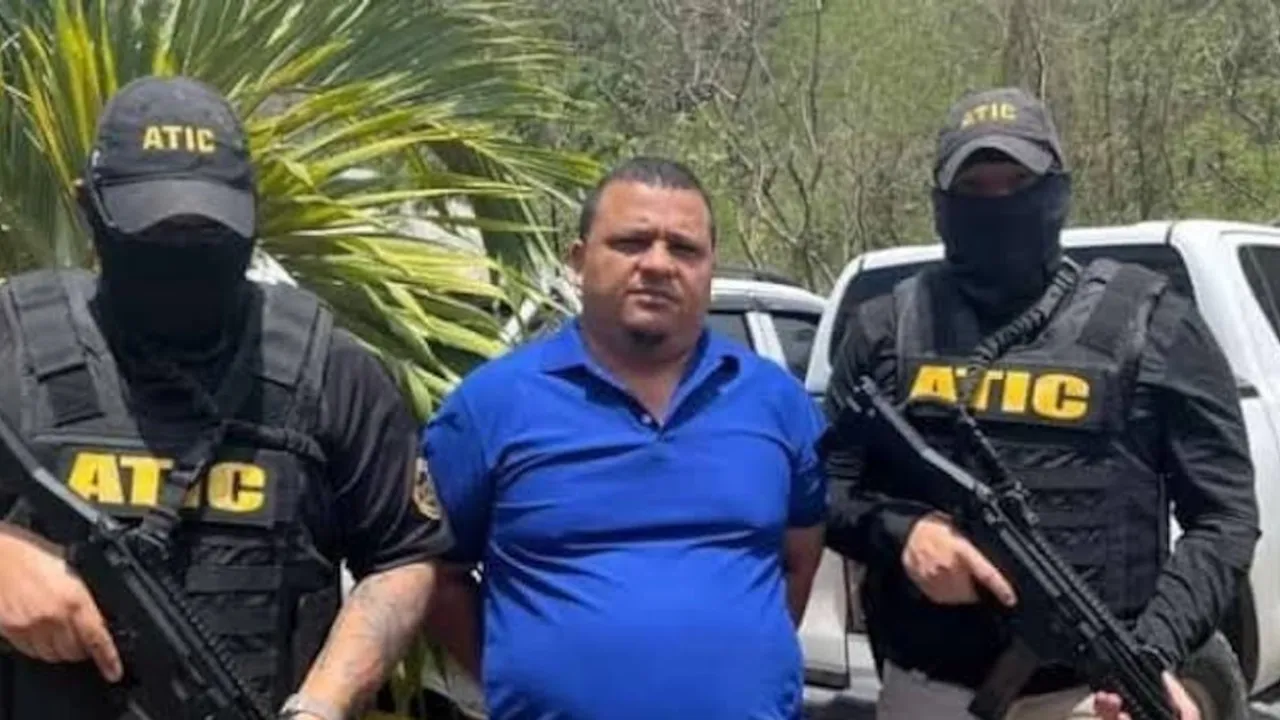 Hombre con remera azul custodiado por dos agentes de seguridad con pasamontañas, chalecos de ATIC y armas largas.