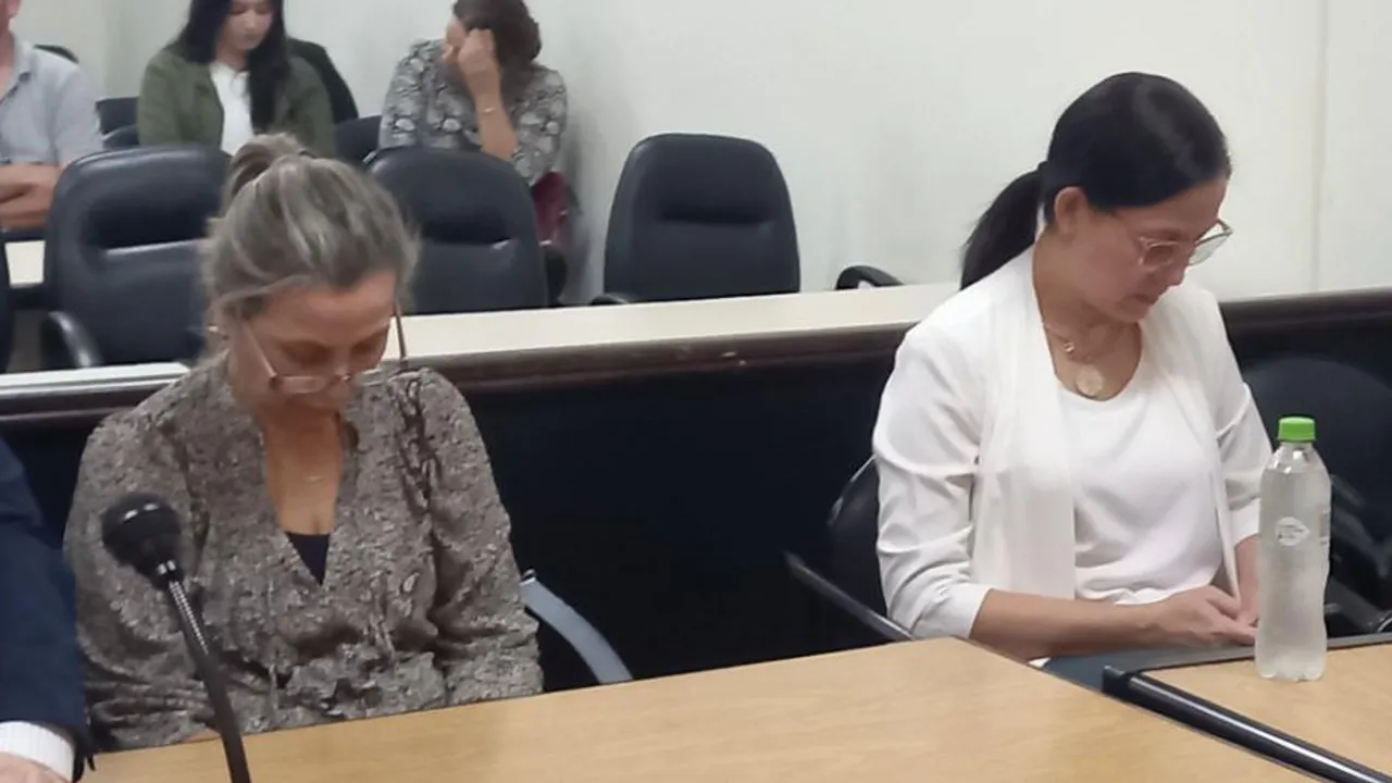 Dos mujeres con anteojos sentadas en un escritorio frente a micrófonos en una sala de audiencia judicial.