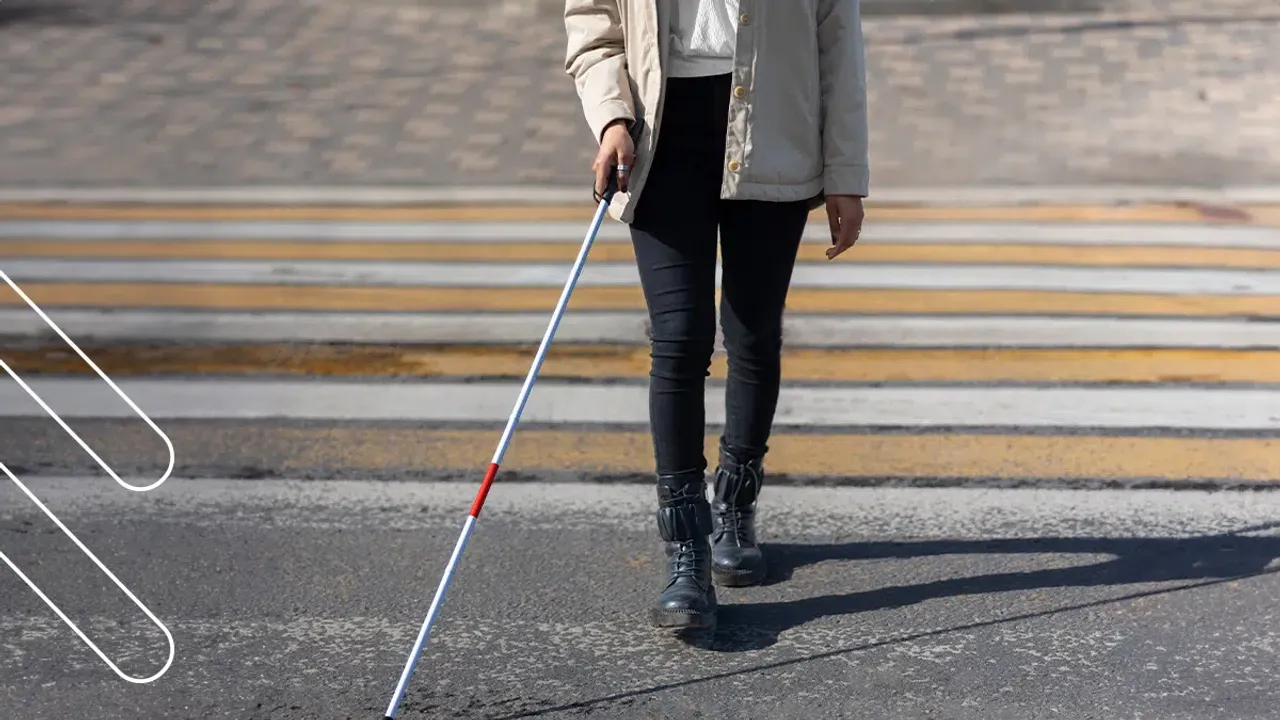 Persona con ropa oscura y bastón blanco cruzando la calle sobre un paso peatonal pintado con franjas amarillas.