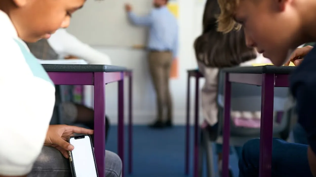 Un alumno sostiene un teléfono móvil oculto bajo su pupitre escolar mientras el docente enseña al fondo del aula.