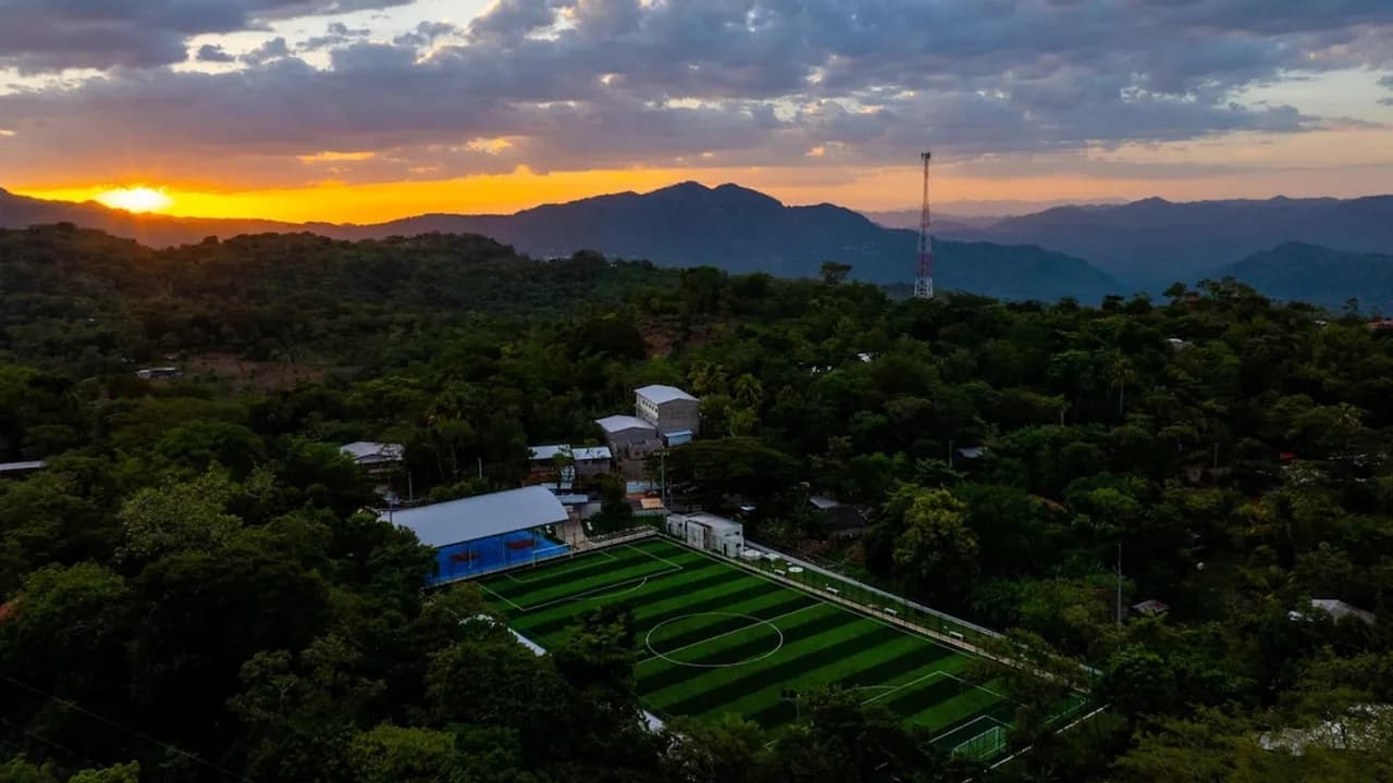 Vista aérea de una cancha sintética y un polideportivo techado rodeados de cerros durante el atardecer.