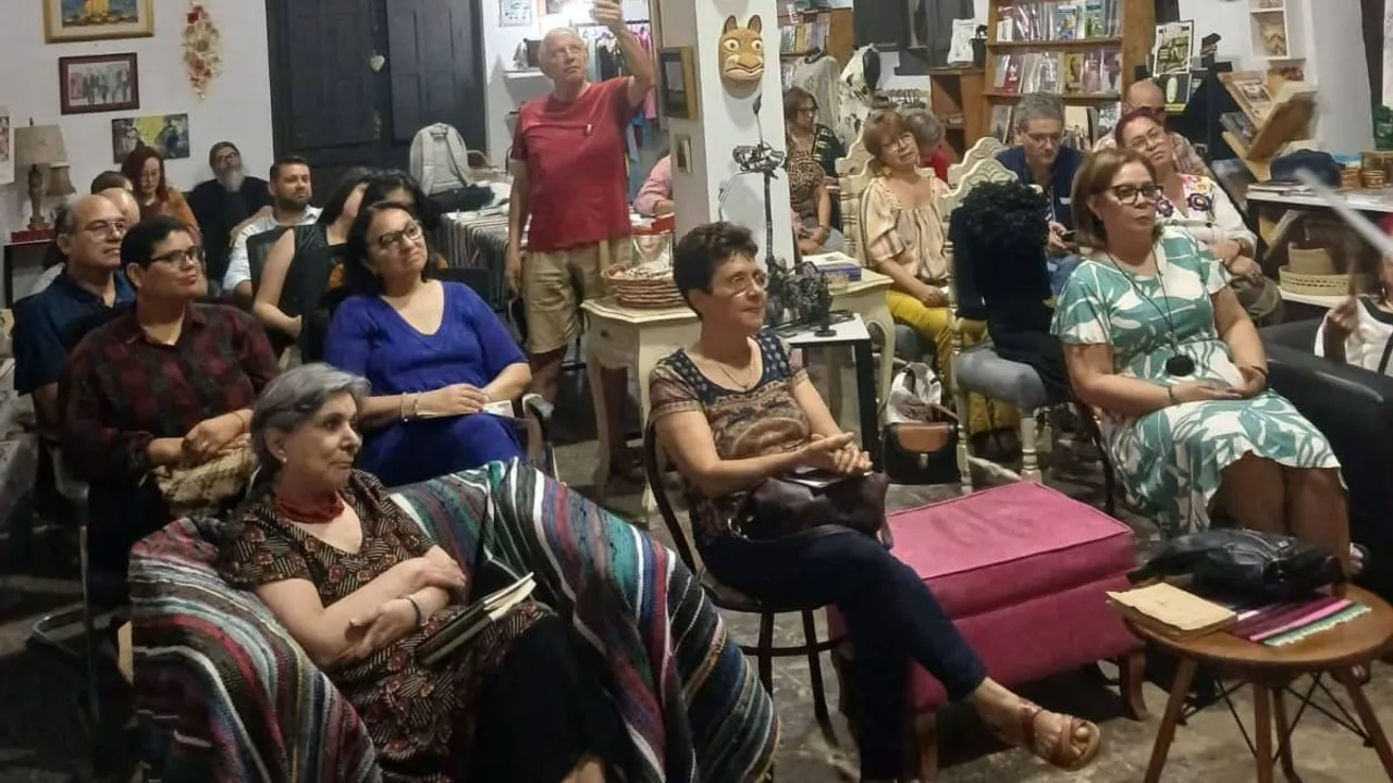Público sentado en una sala cultural con libros y cuadros, prestando atención a una presentación literaria.