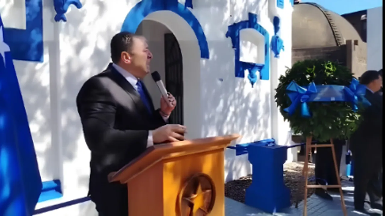 Hombre de traje oscuro habla desde un podio de madera frente a una pared blanca y azul con un micrófono en la mano.