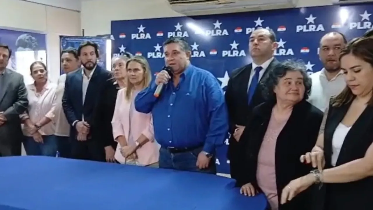 Un hombre habla por micrófono frente a una mesa con varias personas de pie ante un fondo con el logo partidario del PLRA.