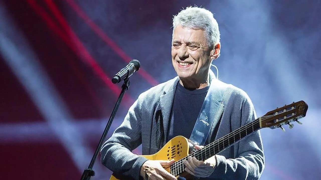 El músico brasileño Chico Buarque sonríe sobre el escenario mientras sostiene su guitarra acústica frente al micrófono.